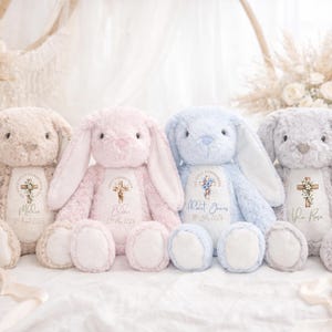 May include: Four plush stuffed bunnies in beige, pink, blue, and gray. Each bunny features a white chest patch printed with a cross, floral accents, and personalized text including names and dates for a First Holy Communion celebration.