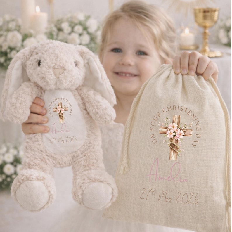 Op de afbeelding: Een beige pluche konijn en een stoffen zak met de tekst "On Your Christening Day". Het konijn en de zak zijn gepersonaliseerd met de naam "Amelia" en de datum "27 mei 2026". De zak heeft een kruisontwerp met bloemaccenten.