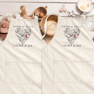 May include: Two white aprons featuring a black and white map design in the shape of a heart. The map has a red pin in the centre. The text "Where it began" and "Laurie & Joe" is printed on each apron.