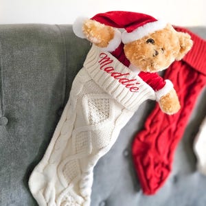 以下が含まれることがあります： クリーム色のケーブル編みのクリスマスストッキング。赤い刺繍で「Maddie」とパーソナライズされています。サンタ帽をかぶったテディベアが覗いています。背景には赤いストッキングがあります。