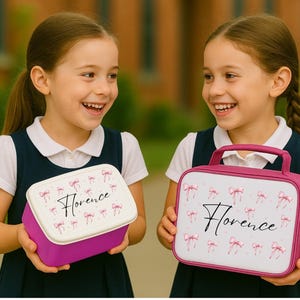 May include: Two girls holding personalized lunchboxes. One is a rectangular box with a white lid, the other is a pink handled bag. Both have a white background with pink bows and the name "Florence" in script.