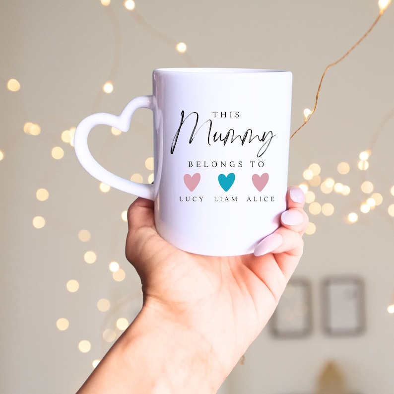 May include: White ceramic mug with a heart-shaped handle. The mug has black text that reads "This Mummy Belongs To" with three hearts underneath. The hearts are pink, teal, and pink, with the names "Lucy", "Liam", and "Alice" written underneath.