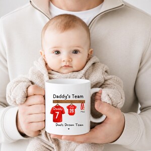 Op de afbeelding: Een witte keramische mok met een afbeelding van twee rode sportshirts die aan een houten rek hangen. De tekst luidt Daddy's Team boven de shirts en Dad's Dream Team eronder. E&eacute;n shirt is gelabeld met Daddy 7 en het andere met Atlas 26.
