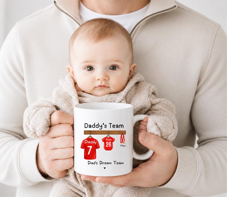 Op de afbeelding: Een witte keramische mok met een afbeelding van twee rode sportshirts die aan een houten rek hangen. De tekst luidt Daddy's Team boven de shirts en Dad's Dream Team eronder. E&eacute;n shirt is gelabeld met Daddy 7 en het andere met Atlas 26.