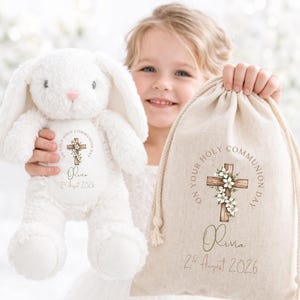 May include: A white plush bunny toy and a beige drawstring canvas bag, both featuring a printed design of a wooden cross adorned with white flowers, the text "ON YOUR HOLY COMMUNION DAY," the name "Olivia," and the date "2nd August 2026."