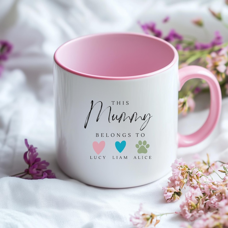 May include: White ceramic mug with a pink rim and handle. The mug has black text that reads "This Mummy Belongs To" with three icons below: a pink heart, a blue heart, and a paw print. The icons are labeled "Lucy", "Liam", and "Alice".