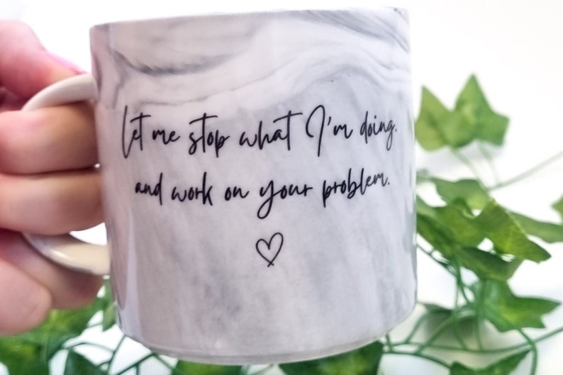 K&ouml;nnte beinhalten: Wei&szlig;e Keramiktasse mit Marmormuster und schwarzem Text, der "Lass mich aufh&ouml;ren, was ich tue, und an deinem Problem arbeiten." Ein kleines Herz befindet sich unter dem Text.