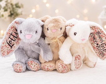 Könnte beinhalten: Drei Plüschhasen in Grau, Beige und Weiß. Der graue Hase hat den Namen Lottie auf sein geblümtes Ohr gestickt. Der weiße Hase trägt die Aufschrift Flower Girl und 00-06-2028 auf seinem Ohr. Alle haben rosa Nasen und Schleifen.
