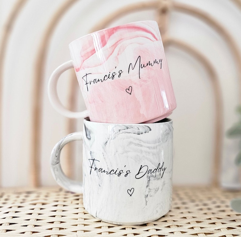 May include: Two ceramic mugs with marbleized designs. The top mug is pink with the text "Francis's Mummy" and a small heart. The bottom mug is gray with the text "Francis's Daddy" and a small heart.