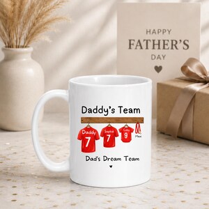 Op de afbeelding: Een witte keramische mok met de tekst Daddy's Team en Dad's Dream Team. Het ontwerp toont drie rode sportshirts hangend aan een houten rek, gelabeld met Daddy, Sophie en Coby met de nummers 7, 7 en 9, plus een rode hondenriem gelabeld Maxi.