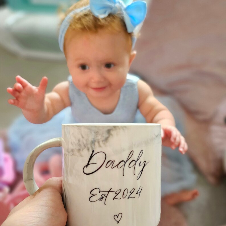 May include: A white ceramic mug with a marbleized design. The mug has black text that reads "Daddy Est 2024" with a small heart symbol below the text. A baby in a blue dress and a blue bow is looking at the mug.