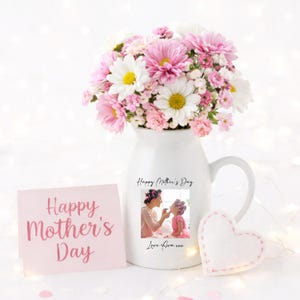 May include: A white ceramic pitcher filled with pink and white flowers. A pink card with "Happy Mother's Day" is next to the pitcher. The pitcher has a photo and text that says "Happy Mother's Day" and "Love Ava xxx". A white felt heart is also present.