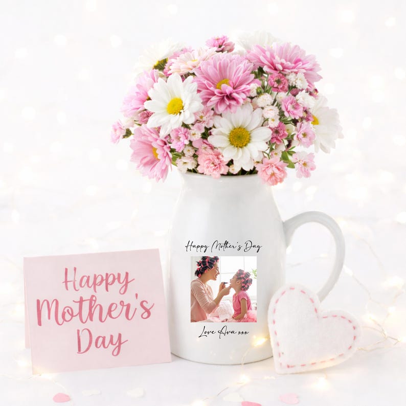 May include: A white ceramic pitcher filled with pink and white flowers. A pink card with "Happy Mother's Day" is next to the pitcher. The pitcher has a photo and text that says "Happy Mother's Day" and "Love Ava xxx". A white felt heart is also present.
