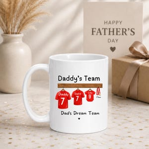 Op de afbeelding: Een witte keramische mok met de tekst Daddy's Team en Dad's Dream Team. Het ontwerp toont drie rode sportshirts hangend aan een houten rek, gelabeld met Daddy, Sophie en Coby met de nummers 7, 7 en 9, plus een rode hondenriem gelabeld Maxi.