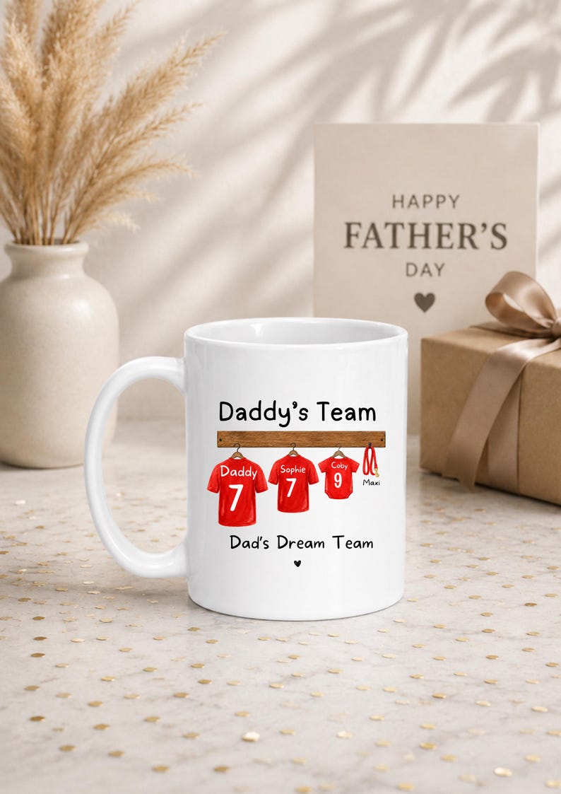 Op de afbeelding: Een witte keramische mok met de tekst Daddy's Team en Dad's Dream Team. Het ontwerp toont drie rode sportshirts hangend aan een houten rek, gelabeld met Daddy, Sophie en Coby met de nummers 7, 7 en 9, plus een rode hondenriem gelabeld Maxi.