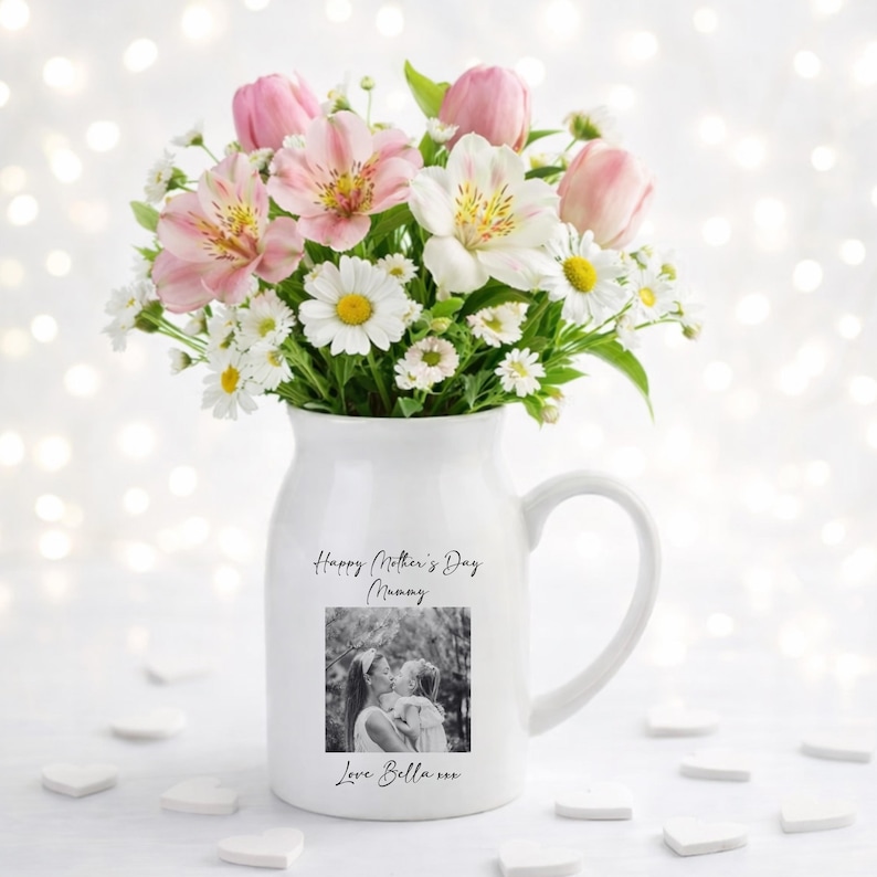May include: A white ceramic mug with a black handle and the text "But First, Coffee" in a bold, black font. The mug is set against a blurred background. The mug appears to be approximately 10 cm tall.