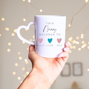 May include: White ceramic mug with a heart-shaped handle. The mug has the text "This Nanny Belongs To" with three hearts and the names "Milly", "Liam", and "Mia" underneath.