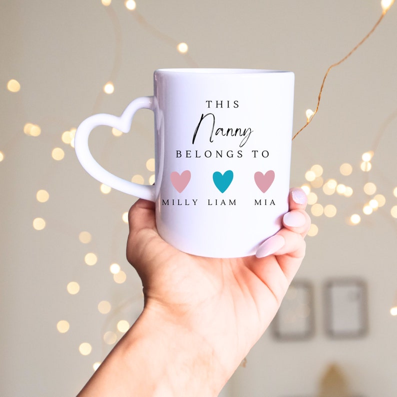 May include: White ceramic mug with a heart-shaped handle. The mug has the text "This Nanny Belongs To" with three hearts and the names "Milly", "Liam", and "Mia" underneath.