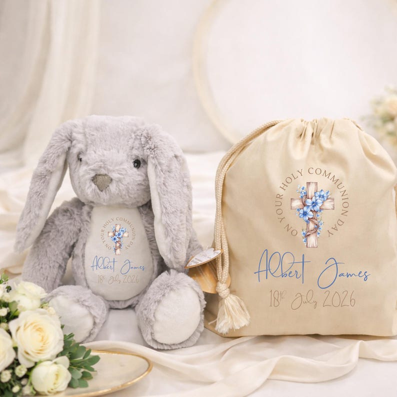 Op de afbeelding: Een grijze pluche konijn en een beige trekkoordtas. De tas en het konijn hebben een kruisontwerp met blauwe bloemen en de tekst "YOUR HOLY COMMUNION DAY". De tas en het konijn zijn gepersonaliseerd met de naam "Albert James" en de datum "18 juli 2026".