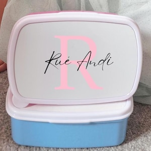 May include: A light blue and white lunchbox with a pink lid. The lid features a large pink letter "R" and the name "Rue Andi" in black script. The lunchbox is rectangular and has a white rim.