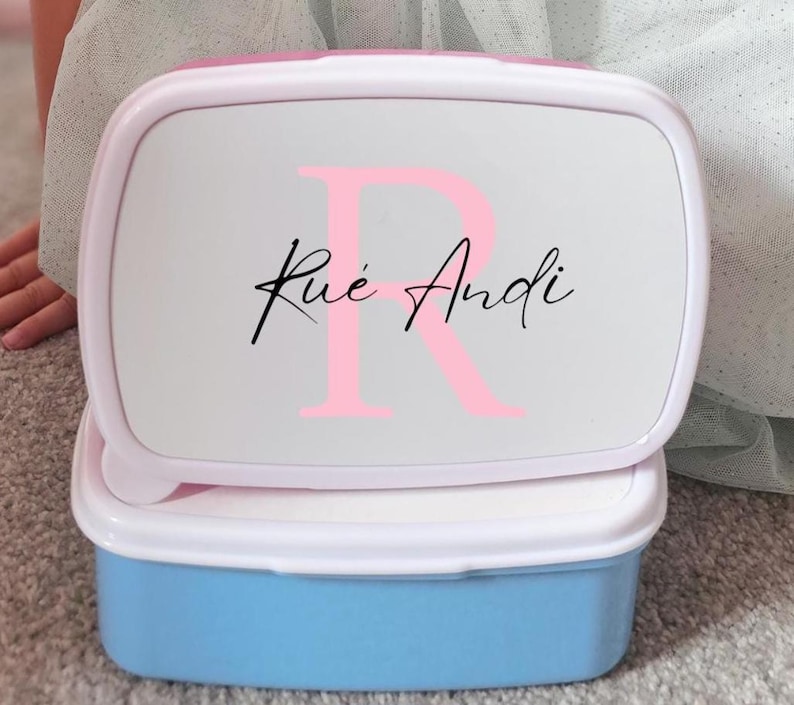 May include: A light blue and white lunchbox with a pink lid. The lid features a large pink letter "R" and the name "Rue Andi" in black script. The lunchbox is rectangular and has a white rim.