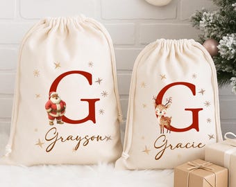 Puede incluir: Dos bolsas con cordón de color crema con diseños navideños. Una presenta una ilustración de Papá Noel y el nombre "Grayson", la otra una ilustración de ciervo y el nombre "Gracie". Ambas tienen una gran letra "G" roja y estrellas.