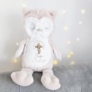 Op de afbeelding: Een zachtroze en witte uilknuffel met een kruisontwerp met bloemaccenten en de tekst "ON YOUR CHRISTENING DAY" en "Jasper 2nd August 2025". De uil heeft gesloten ogen. Hij zit op een grijze ondergrond.