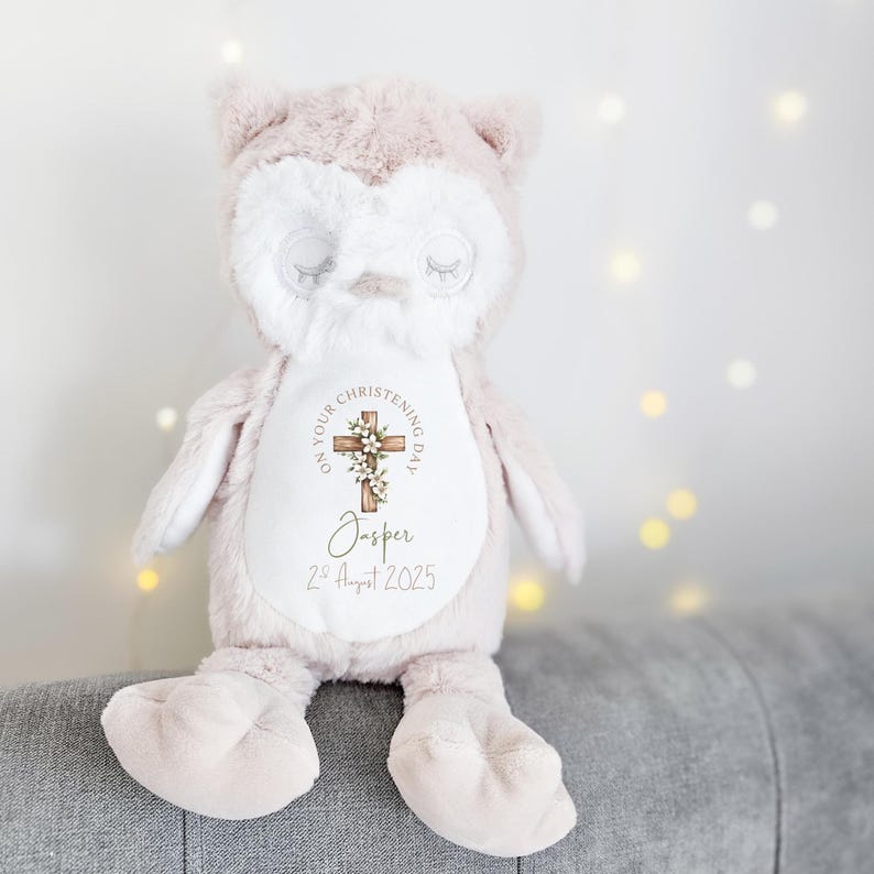 Op de afbeelding: Een zachtroze en witte uilknuffel met een kruisontwerp met bloemaccenten en de tekst "ON YOUR CHRISTENING DAY" en "Jasper 2nd August 2025". De uil heeft gesloten ogen. Hij zit op een grijze ondergrond.