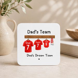 Op de afbeelding: Een vierkante witte onderzetter met de tekst Dad's Team en Dad's Dream Team, voorzien van een afbeelding van drie rode shirts aan een houten rek, gelabeld met Dad, Sophie en Coby met de nummers 7, 7 en 9, en een rode riem genaamd Maxi.