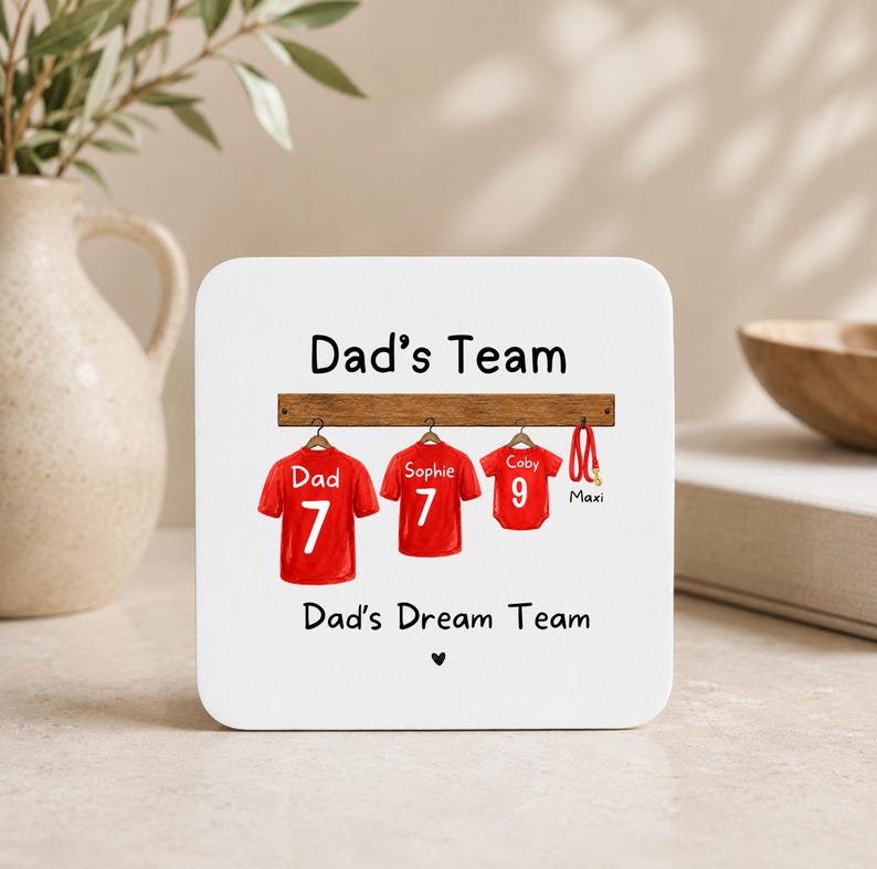 Op de afbeelding: Een vierkante witte onderzetter met de tekst Dad's Team en Dad's Dream Team, voorzien van een afbeelding van drie rode shirts aan een houten rek, gelabeld met Dad, Sophie en Coby met de nummers 7, 7 en 9, en een rode riem genaamd Maxi.