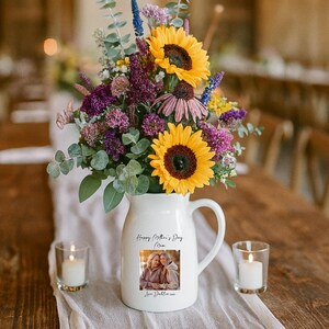 May include: A white ceramic pitcher with a handle, filled with a vibrant floral arrangement including sunflowers and purple flowers. The pitcher has a photo and text that reads "Happy Mother's Day Mom". Two lit candles sit on either side.