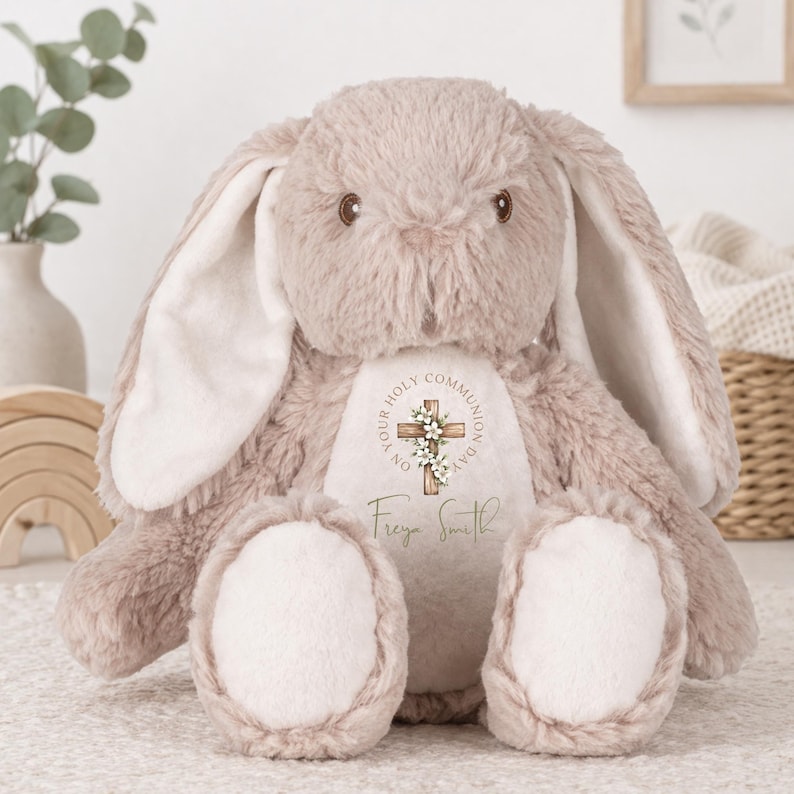May include: A plush beige bunny with long floppy ears and a cream-colored belly patch. The belly features a printed wooden cross adorned with white flowers, the text "ON YOUR HOLY COMMUNION DAY," and the personalized name "Freya Smith" in script.