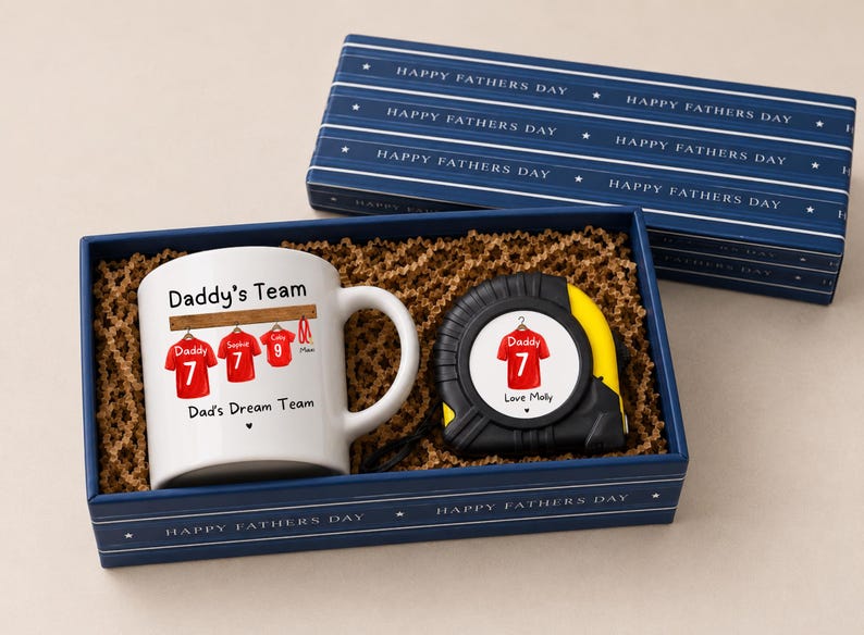 Op de afbeelding: Een witte keramische mok en een zwart-geel uittrekbaar meetlint in een blauwe geschenkdoos. De mok bevat de tekst Daddy's Team en Dad's Dream Team met rode shirtillustraties. Het meetlint heeft een afbeelding van een rood shirt.