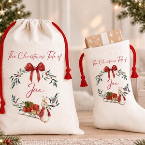 May include: A cream-coloured drawstring gift bag and a matching Christmas stocking, both featuring the text The Christmas Tale of Jax, a red bow, a green wreath, and a rabbit illustration with gift boxes. The items have red cord drawstrings.