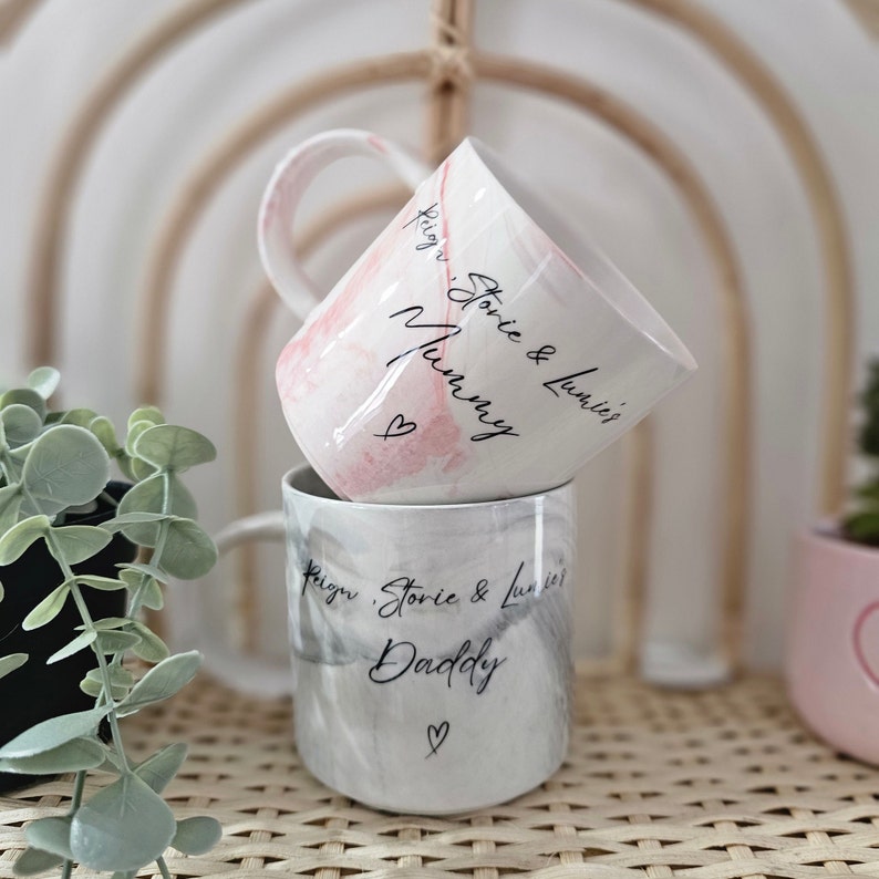 May include: Two ceramic mugs with marbleized designs. The top mug is pink and says "Reign, Storie & Lumi's Mummy." The bottom mug is gray and says "Reign, Storie & Lumi's Daddy."