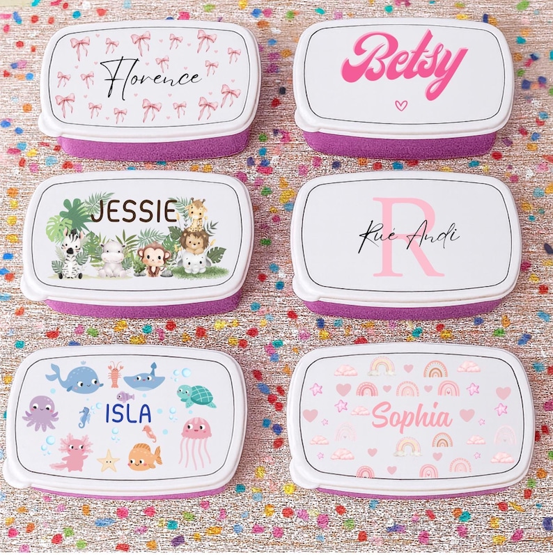 May include: Six personalized lunch boxes with white lids and purple bases. The lids feature various designs, including names like "Florence" and "Betsy," along with decorative elements such as bows, hearts, and animal illustrations. The lunch boxes are rectangular.