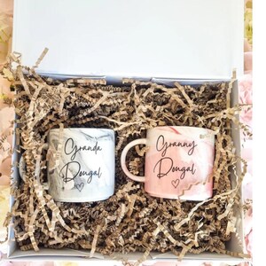 May include: A white gift box with brown shredded paper inside. Two white ceramic mugs with marbleized designs are inside the box. One mug says "Granda Dougal" and the other says "Granny Dougal".