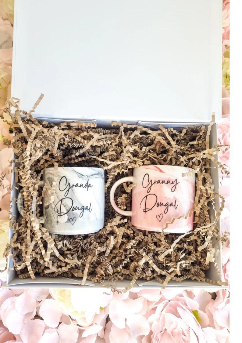 May include: A white gift box with brown shredded paper inside. Two white ceramic mugs with marbleized designs are inside the box. One mug says "Granda Dougal" and the other says "Granny Dougal".