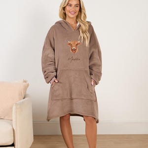 May include: A brown fleece hooded blanket with a large pocket in the front. The blanket features a printed image of a Highland cow and the name "Maddie" in cursive.