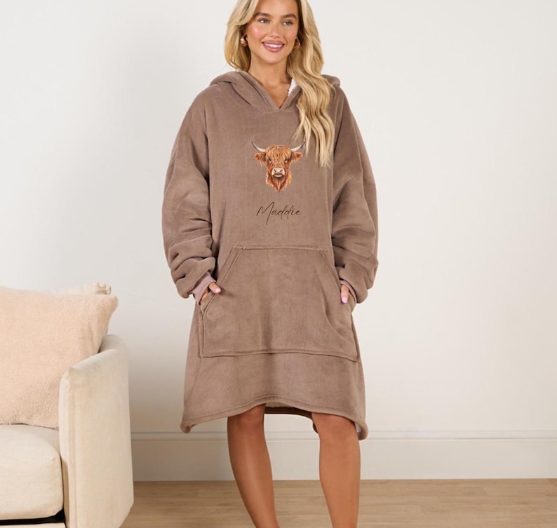 May include: A brown fleece hooded blanket with a large pocket in the front. The blanket features a printed image of a Highland cow and the name "Maddie" in cursive.
