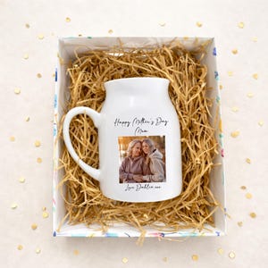 May include: A white ceramic mug with a large handle, featuring a photo of two women and the text "Happy Mother's Day Mum" and "Love Dahlia xxx". The mug is presented in a decorative box filled with straw.