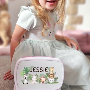 May include: A personalized lunchbox with a pink and white lid and a blue base. The lid features the name "Jessie" and a jungle animal design including a zebra, monkey, and giraffe. The lunchbox is open, revealing the interior.