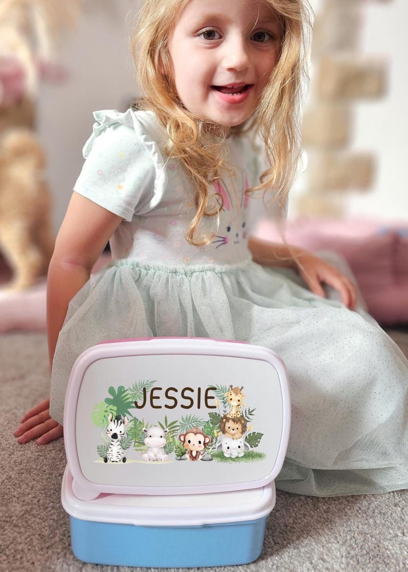 May include: A personalized lunchbox with a pink and white lid and a blue base. The lid features the name "Jessie" and a jungle animal design including a zebra, monkey, and giraffe. The lunchbox is open, revealing the interior.