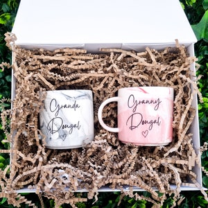 K&ouml;nnte beinhalten: Zwei Keramikbecher mit marmorierten Designs. Ein Becher ist wei&szlig; mit schwarzer Marmorierung und tr&auml;gt die Aufschrift "Granda Dougal" mit einem Herz. Der andere Becher ist rosa mit wei&szlig;er Marmorierung und tr&auml;gt die Aufschrift "Granny Dougal" mit einem Herz. Die Becher befinden sich in einer wei&szlig;en Box mit braunem zerkleinertem Papier.