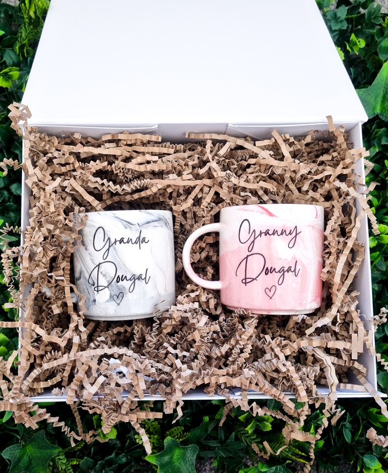 K&ouml;nnte beinhalten: Zwei Keramikbecher mit marmorierten Designs. Ein Becher ist wei&szlig; mit schwarzer Marmorierung und tr&auml;gt die Aufschrift "Granda Dougal" mit einem Herz. Der andere Becher ist rosa mit wei&szlig;er Marmorierung und tr&auml;gt die Aufschrift "Granny Dougal" mit einem Herz. Die Becher befinden sich in einer wei&szlig;en Box mit braunem zerkleinertem Papier.