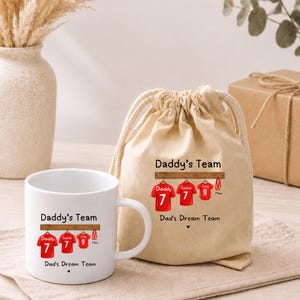 Op de afbeelding: Een witte keramische mok en een beige stoffen tas met trekkoord, beide voorzien van een afbeelding van drie rode sportshirts aan een houten rek met de tekst Daddy's Team en Dad's Dream Team in het zwart gedrukt en een klein hartje.
