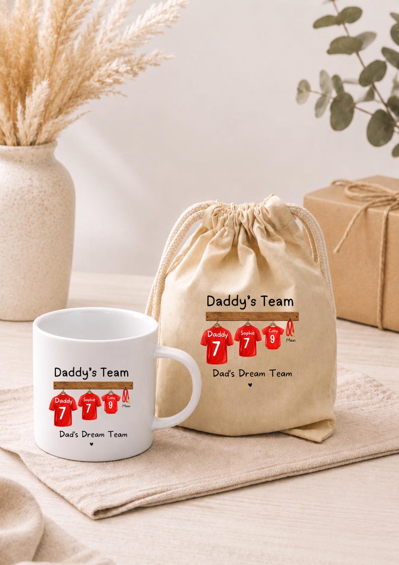 Op de afbeelding: Een witte keramische mok en een beige stoffen tas met trekkoord, beide voorzien van een afbeelding van drie rode sportshirts aan een houten rek met de tekst Daddy's Team en Dad's Dream Team in het zwart gedrukt en een klein hartje.