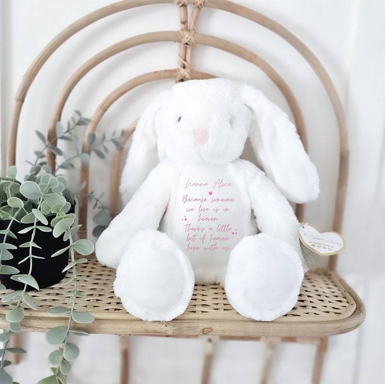 May include: White plush bunny with long ears, sitting on a woven chair. The bunny has pink embroidered details and a message that reads "hanna Alice Because someone we love is in heaven There's a little bit of heaven here with us".