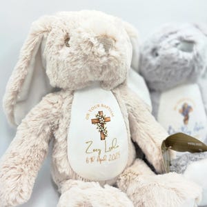 Op de afbeelding: Een zacht, beige konijn met lange oren en een witte buik. De buik heeft een kruisontwerp met de tekst "ON YOUR BAPTISM", een naam en een datum. Een tweede grijs konijn is gedeeltelijk zichtbaar op de achtergrond.