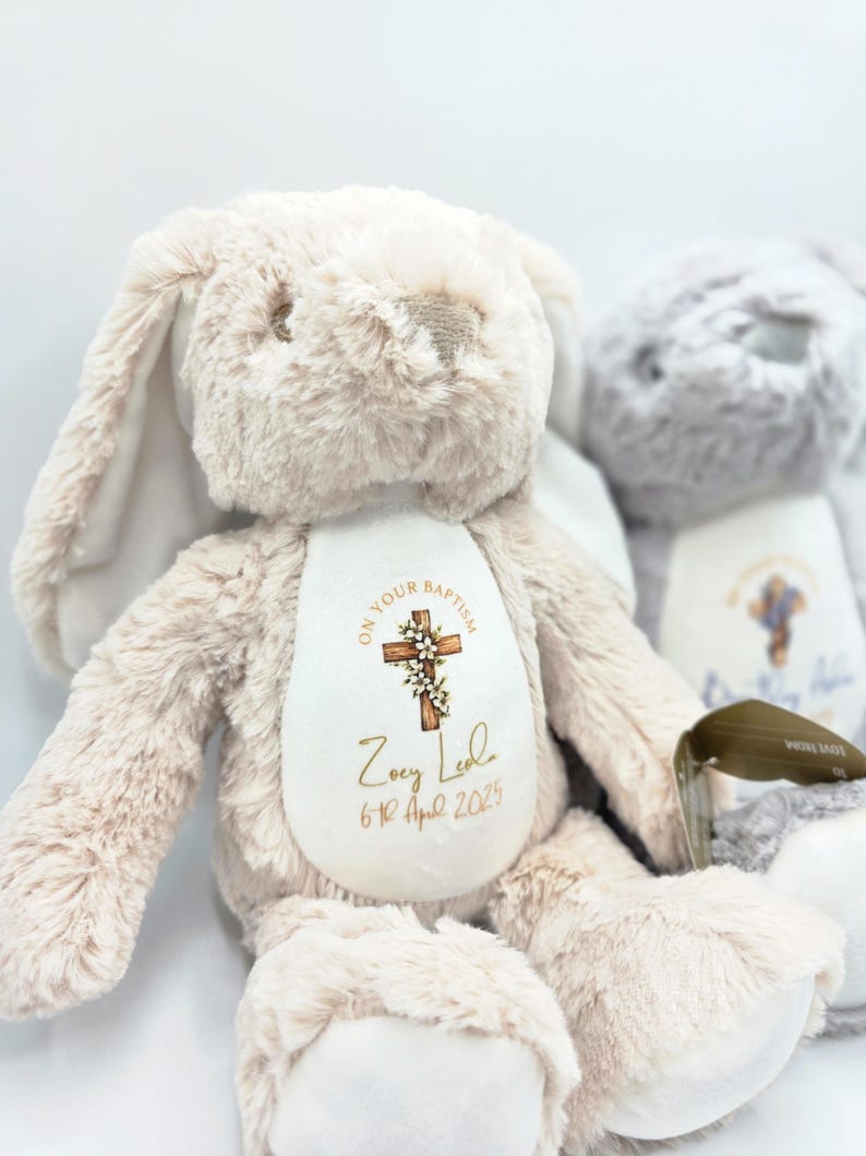 Op de afbeelding: Een zacht, beige konijn met lange oren en een witte buik. De buik heeft een kruisontwerp met de tekst "ON YOUR BAPTISM", een naam en een datum. Een tweede grijs konijn is gedeeltelijk zichtbaar op de achtergrond.